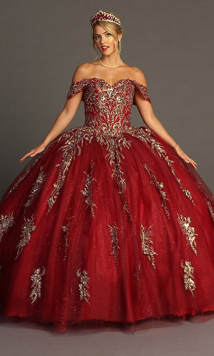 A woman wearing the Quinceanera Dress CHF3084 By Chicas in a rich burgundy shade, featuring off-the-shoulder glitter tulle ballgown layers adorned with intricate gold floral embroidery and paired with a matching tiara, stands against a plain background.